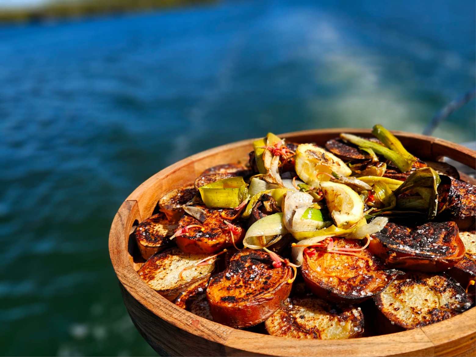Grilled vegetables in a wooden bowl with a blurred blue ocean background.