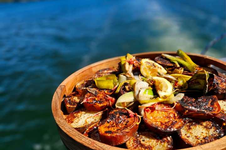 Grilled vegetables in a wooden bowl with a blurred blue ocean background.