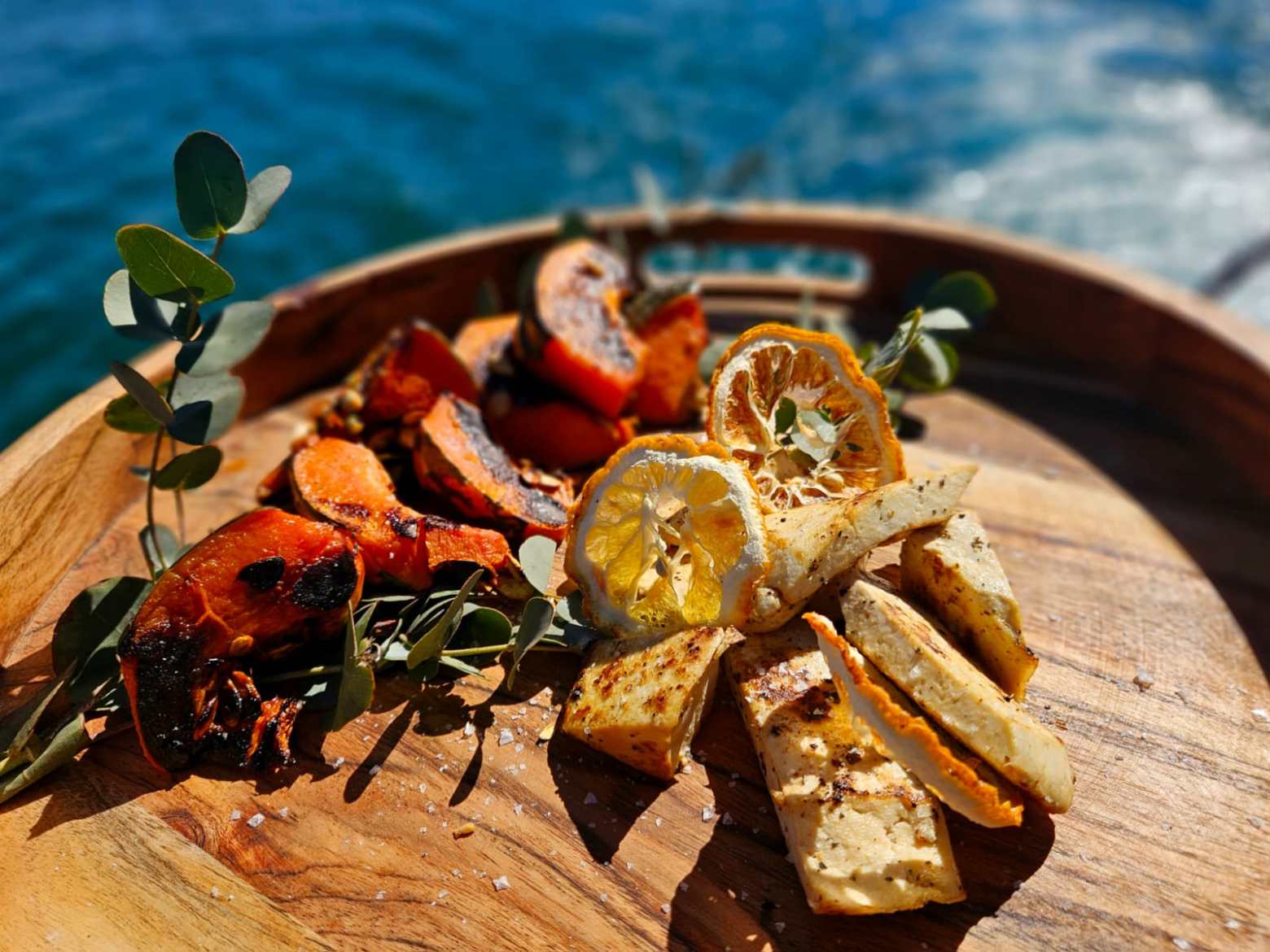 Grilled vegetables and lemon slices on a wooden platter by the sea.