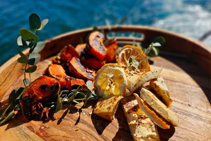 Grilled vegetables and lemon slices on a wooden platter by the sea.