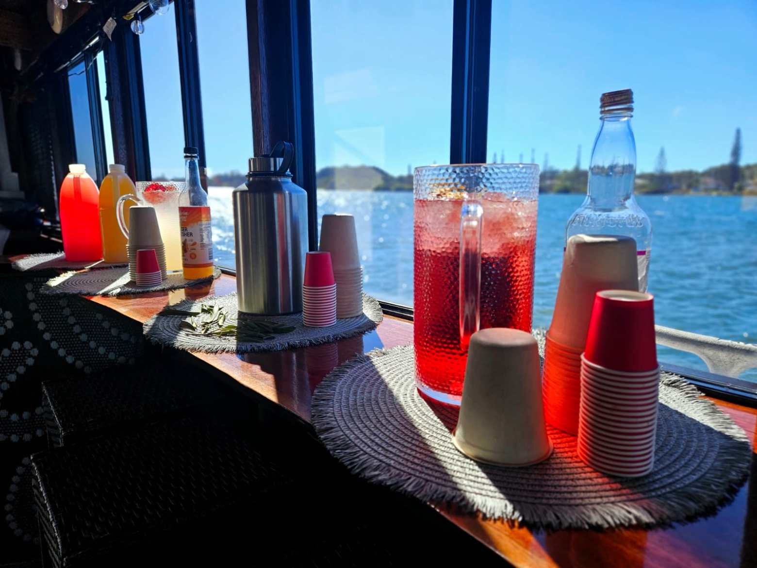 Drink station with red and yellow juices, cups, and a teapot on a boat with water view.