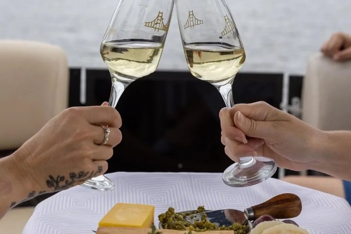 Two hands clink wine glasses over a boat picnic spread on a lake.